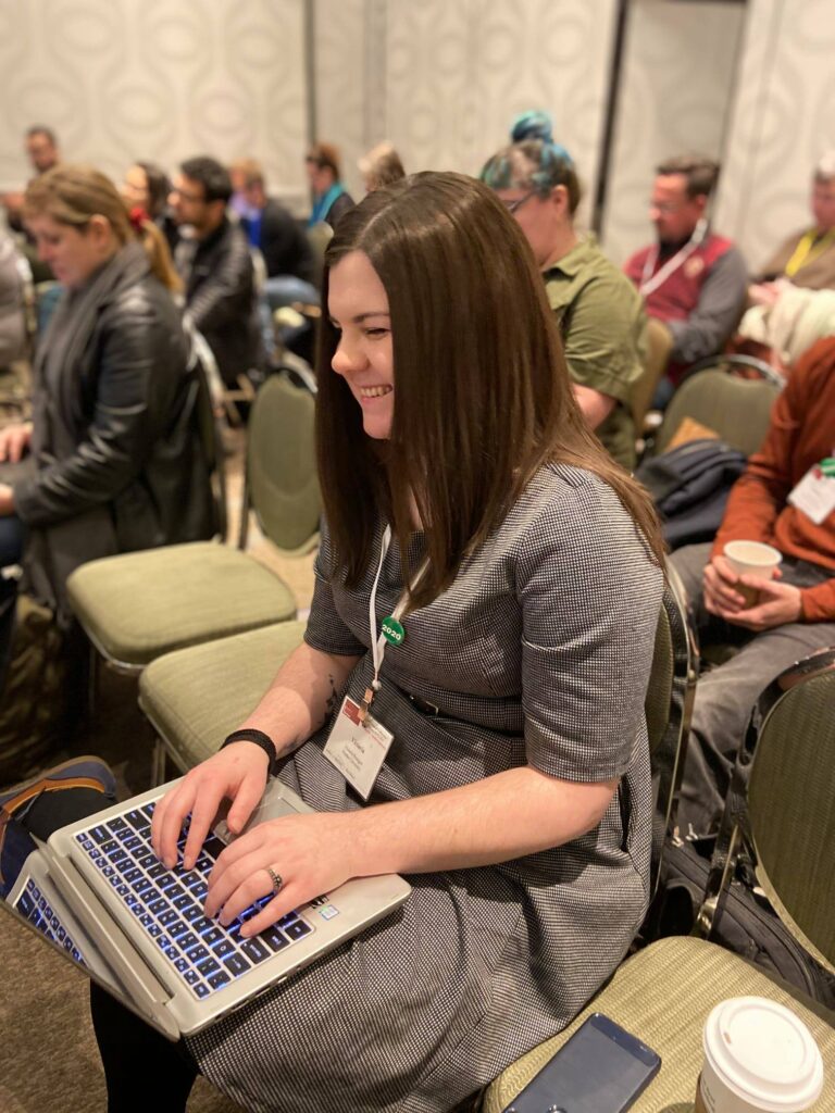 Image of Victoria at a conference, wearing a grey dress with her brown hair straight and hanging loose around her shoulders. She is sitting on a chair and there are conference attendees visible in the background. Her laptop is on her lap and both hands are on the keys. She is laughing and looking away from the camera. 