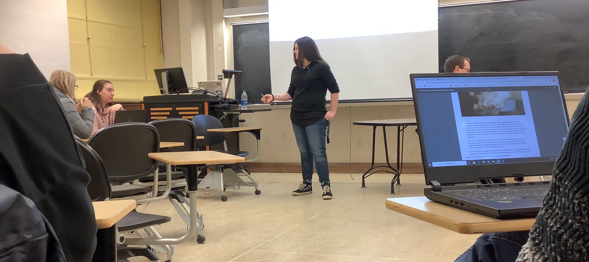 Picture of Victoria Braegger, a woman with long brunette hair, wearing a black tunic sweater, blue jeans, and high tops standing in the center of a classroom. She is listening to a student's answer to a question with one arm extended and the other resting on her pocket. There is a projector screen and chalkboard behind her.