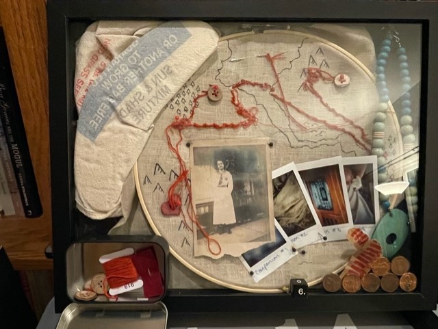 A shadow box for Something Wild. It is chaotic and messy. The map is completed in red, messy stitches and threads along the route. The seedbag for the game is included, but the contents are displayed in the shadowbox: a black and white image of a young woman in her early 20s, and three blurry polaroids are visible. 